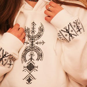 Peut inclure: Sweat-shirt blanc avec un motif géométrique noir sur le devant et les manches. Le motif présente un motif répétitif de triangles, de cercles et de carrés.