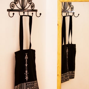 May include: Two black tote bags with white geometric patterns hang on a black metal hook rack with a decorative scroll design. The bags are hanging on the hooks in a white room with a mirror in the background.