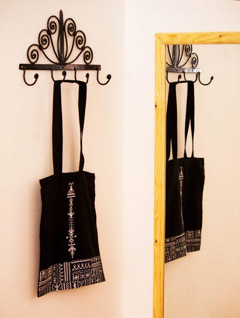 May include: Two black tote bags with white geometric patterns hang on a black metal hook rack with a decorative scroll design. The bags are hanging on the hooks in a white room with a mirror in the background.