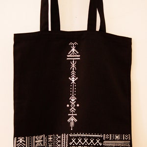 May include: Black canvas tote bag with white geometric designs. The bag has a white design of a vertical arrow with geometric shapes and a white border of geometric patterns.