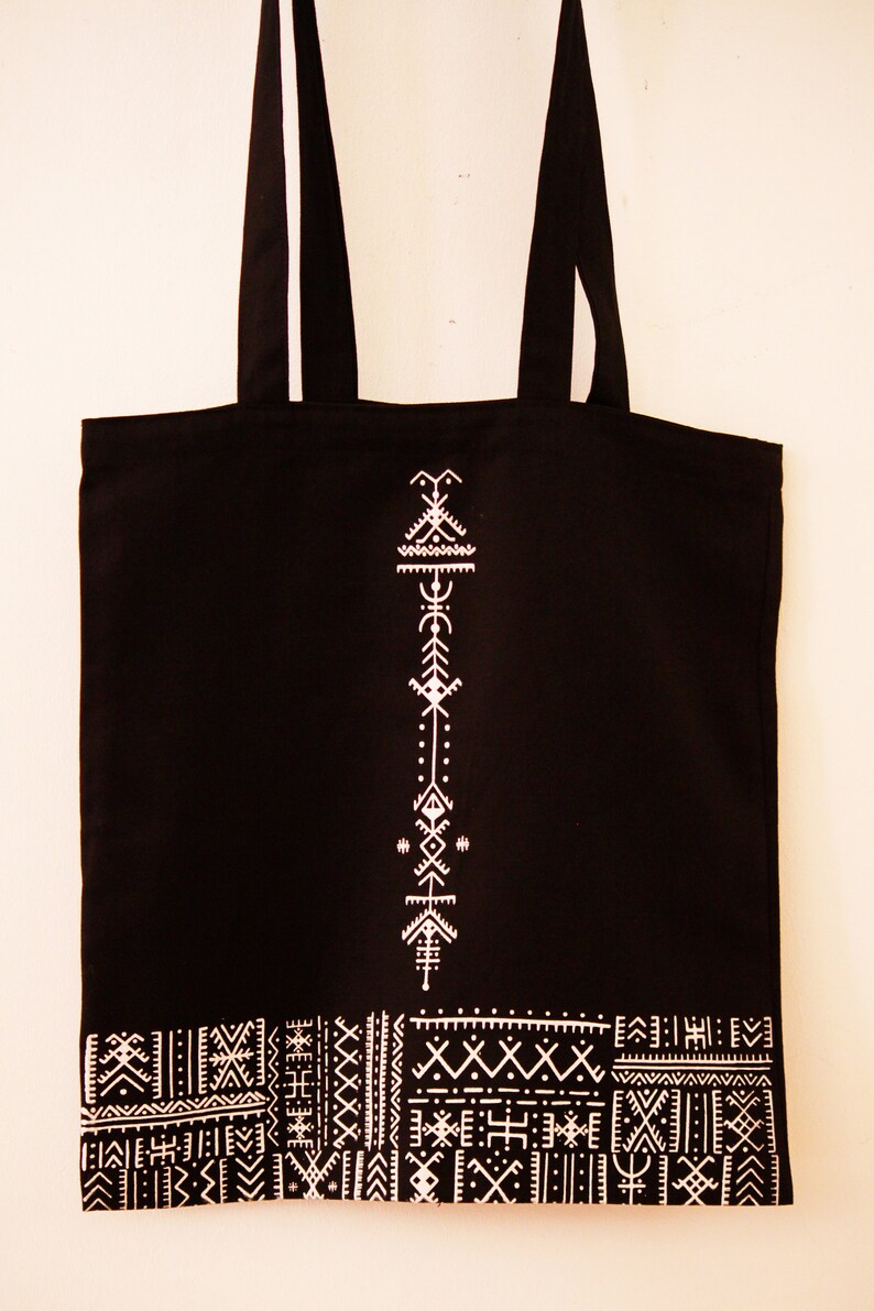May include: Black canvas tote bag with white geometric designs. The bag has a white design of a vertical arrow with geometric shapes and a white border of geometric patterns.