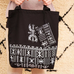 May include: A black tote bag with white geometric patterns and a smaller matching pouch. The bag has a black handle and is held by a person.