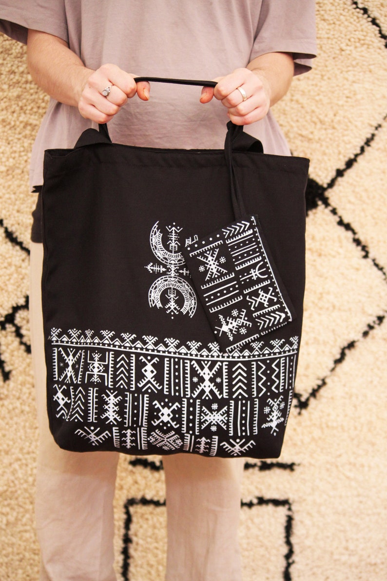 May include: A black tote bag with white geometric patterns and a smaller matching pouch. The bag has a black handle and is held by a person.
