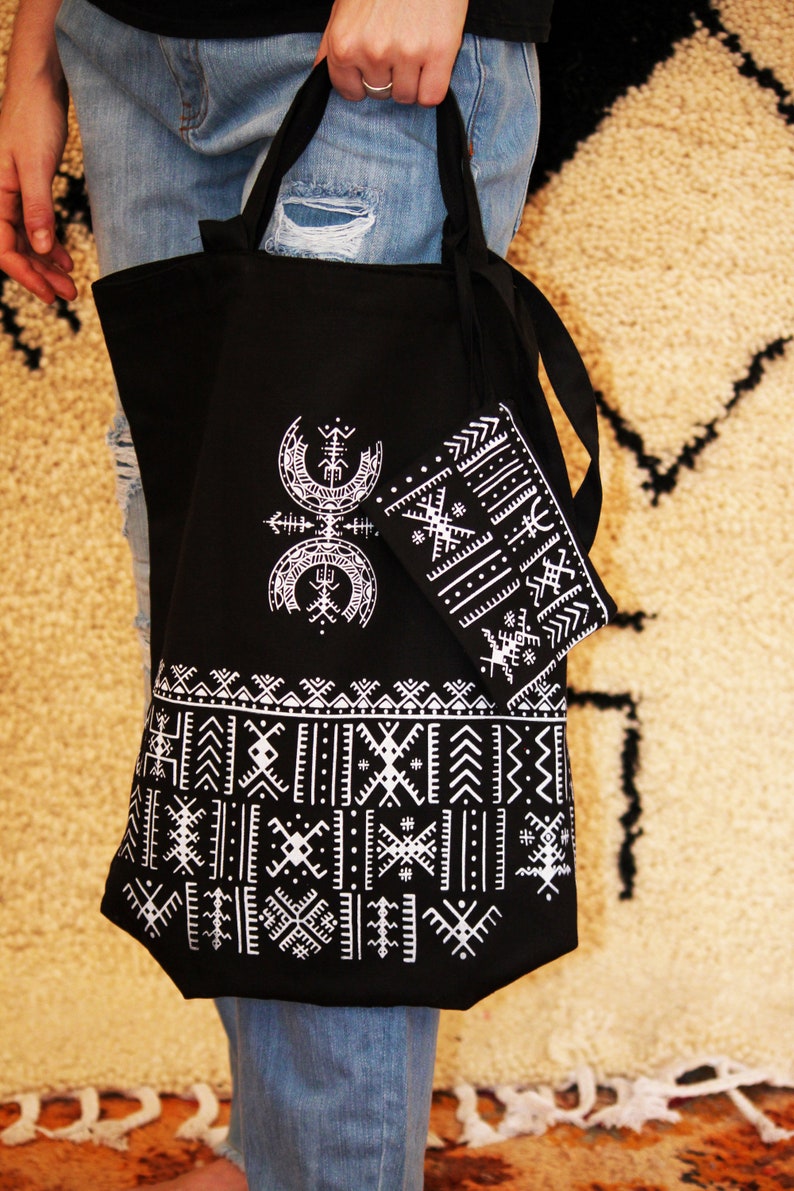 May include: A black tote bag with white geometric patterns and a smaller matching pouch. The bag has two long straps and is made of canvas.