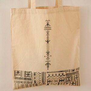 May include: A beige canvas tote bag with black geometric patterns printed on the front. The design features a vertical line of abstract symbols and a horizontal band of repeating patterns.