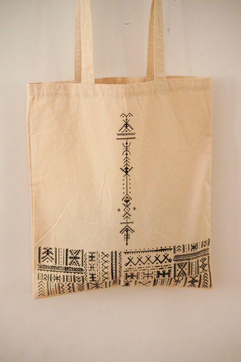 May include: A beige canvas tote bag with black geometric patterns printed on the front. The design features a vertical line of abstract symbols and a horizontal band of repeating patterns.