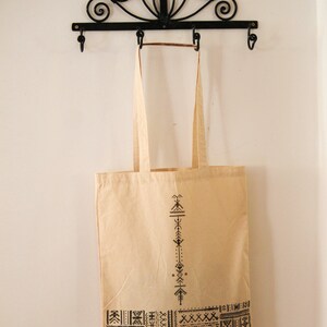 May include: A beige tote bag with black geometric designs. The bag is hanging on a black metal hook with decorative scrolls.