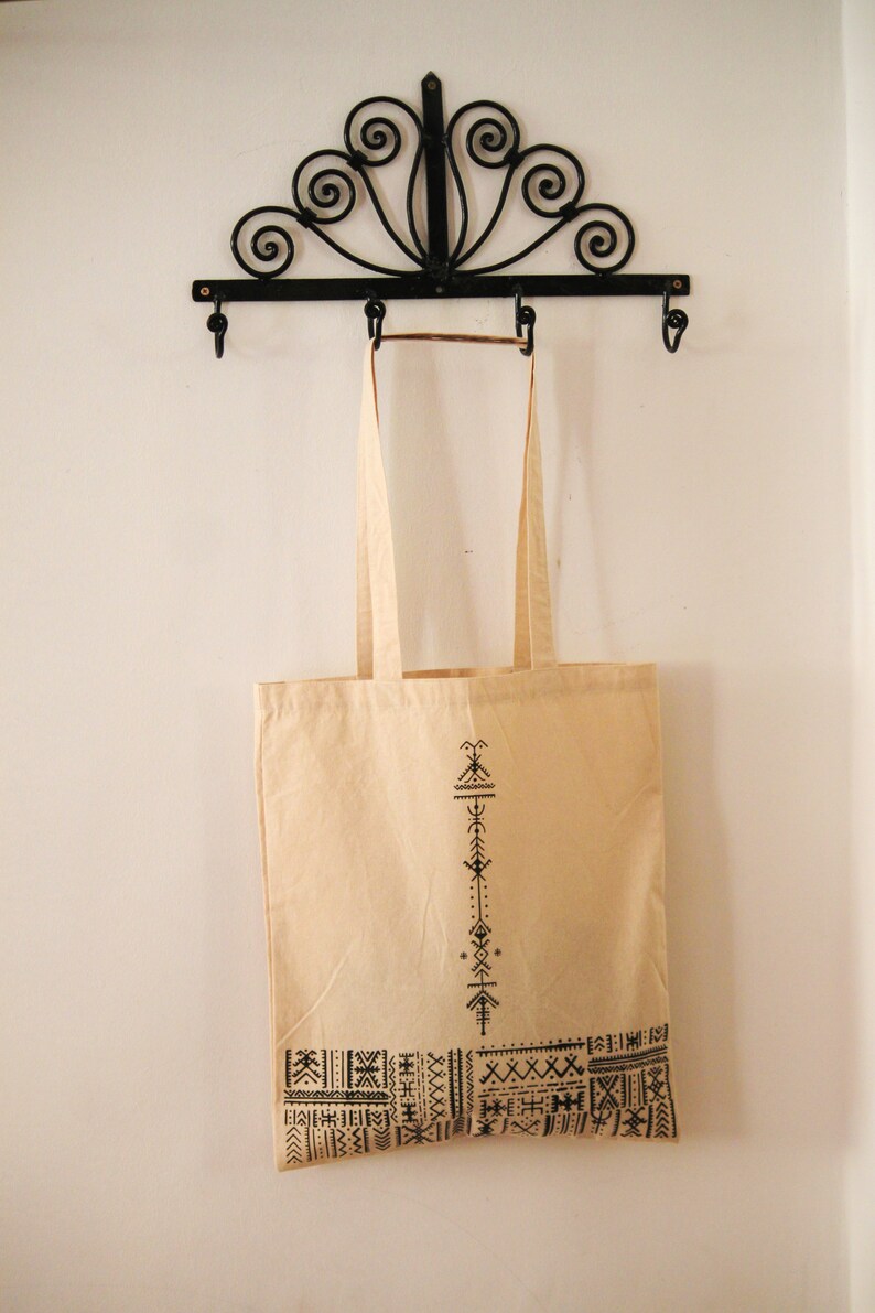 May include: A beige tote bag with black geometric designs. The bag is hanging on a black metal hook with decorative scrolls.