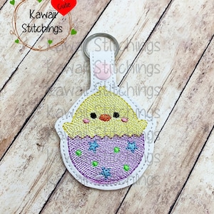 May include: A white keychain with a yellow chick peeking out of a purple egg with blue and green stars. The keychain has a pink button and a silver ring.