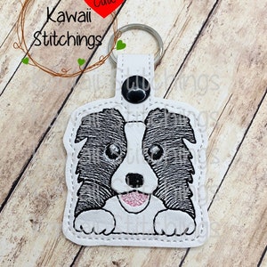 Könnte beinhalten: Ein weißer Leder-Schlüsselanhänger mit einem gestickten Border-Collie-Hundegesicht in Schwarzweiß. Der Schlüsselanhänger hat einen schwarzen Knopf und einen silbernen Schlüsselring. Der Text "Kawaii Stitchings" und "Cute" ist im Hintergrund zu sehen.