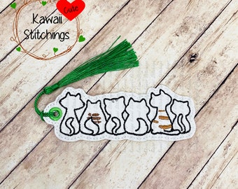 May include: White fabric bookmark with a green tassel. The bookmark features a row of five embroidered cats, with one cat having brown stripes. The bookmark has the text "Kawaii Stitchings" and a heart with the word "Cute" inside.