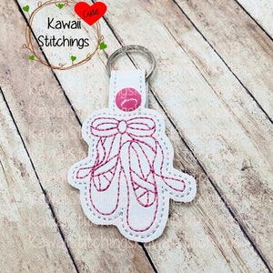 May include: White leather keychain with a pink embroidered design of a pair of ballet shoes with a bow. The text "Kawaii Stitchings" is on the top of the keychain.