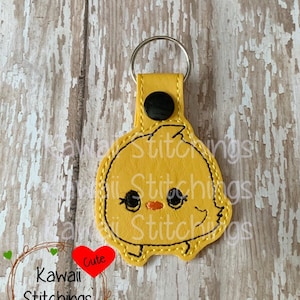 May include: Yellow felt keychain with a cute cartoon chick face. The keychain has a black button and a silver metal ring. The text "Kawaii Stitchings" and "Cute" are visible in the image.
