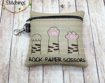 May include: A small, tan zippered pouch with a black zipper and a black clip. The pouch has a brown and black embroidered design of three cat paws with pink paw pads. The text "ROCK PAPER SCISSORS" is embroidered in black on the pouch.