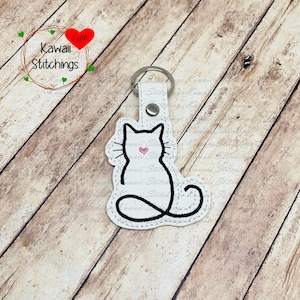 May include: A white cat-shaped keyring with a black outline and a pink heart. The keyring has a silver ring and a white strap. The text "Kawaii Stitchings" is in a circle with a red heart.