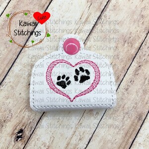 Puede incluir: Llavero de cuero blanco con un corazón rosa y dos huellas de patas negras dentro del corazón. El texto "Kawaii Stitchings" está bordado en la parte superior del llavero.