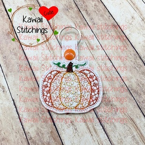 Puede incluir: Un llavero de tela blanca en forma de calabaza con bordados naranja y marrón. El llavero tiene un anillo de metal.