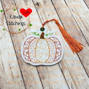 May include: A brown and orange embroidered pumpkin bookmark with a green stem and leaves. The bookmark has a tassel and the text "Kawaii Stitchings" is visible in the background.