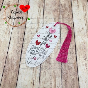 Könnte beinhalten: Ein weißer ovaler Lesezeichen mit einem rosa Quasten. Das Lesezeichen zeigt ein schwarzes gesticktes Design aus zwei Stielen mit Blättern und rosa Herzen.