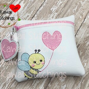 Puede incluir: Una bolsa blanca con cremallera con un globo en forma de corazón rosa y una abeja que sostiene una cuerda atada al globo. La bolsa tiene un dije de corazón rosa unido al tirador de la cremallera. El texto "Kawaii Stitchings" está bordado en la bolsa.
