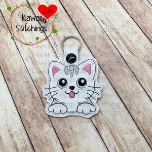 Peut inclure: Porte-clés brodé en forme de chat blanc et gris avec des oreilles roses et un bouton noir. Le porte-clés est sur une surface en bois. Le texte "Cute Kawaii Stitchings" est visible en arrière-plan.