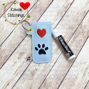 May include: A light blue fabric key chain with a red heart and a black paw print design. The key chain is attached to a small tube of ChapStick labelled "Vanilla Latte".