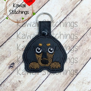 May include: Black and brown dachshund dog face embroidered on a black leather keychain with a silver key ring. The keychain has the text "Kawaii Stitchings" and a red heart with the word "Cute" on it.