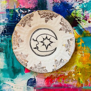 May include: A white decorative plate with a gold rim and floral detailing. The plate has a black illustration of a crescent moon with a closed eye and a star. The background is a colourful abstract painting with pink, blue, and green hues.