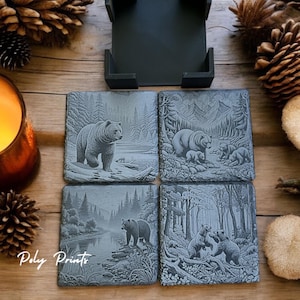May include: Set of four gray slate coasters with a carved design of bears in a forest setting. The coasters are square and have a rustic look. The coasters are arranged in a square shape and are on a wooden surface.