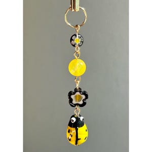 May include: A gold-colored charm with a ladybug design. The charm features a yellow ladybug with black spots, a yellow bead, and two black and white flower-shaped beads. The charm is suspended from a gold-colored ring.