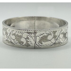 May include: A silver-colored bangle bracelet with an engraved floral design. The bracelet has a hinged clasp and a detailed pattern of leaves and swirls around the band. The metal has a polished finish, reflecting light.