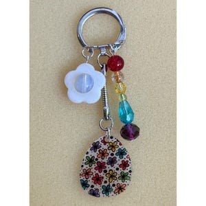 Puede incluir: Un llavero plateado con un dije de flor blanca y un dije en forma de lágrima con patrones florales coloridos. El llavero también presenta una cadena de cuentas con cuentas rojas, amarillas, azules y moradas. Un accesorio decorativo.