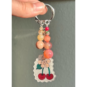 May include: A silver keychain with a charm featuring a cherry blossom design and two red cherries. The keychain also has a strand of colorful beads in shades of orange, yellow, and pink.