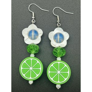 Puede incluir: Pendientes hechos a mano con un diseño de rodaja de lima. Los pendientes tienen una parte superior en forma de flor blanca con un centro azul, cuentas verdes y una rodaja de lima verde y blanca en la parte inferior. Ganchos plateados.