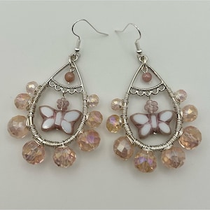 以下が含まれることがあります： シルバーカラーのドロップイヤリングのペア。各イヤリングは、白とピンクの蝶のチャームとピンクと虹色のビーズが付いた、涙の形をしたフレームが特徴です。イヤリングは繊細で手作りの外観をしています。