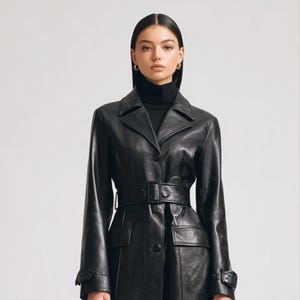 May include: A black leather coat featuring a structured collar, a matching belt with a buckle at the waist, and long sleeves with buckled cuffs. The coat has two front pockets and a button-down front closure, worn over a black turtleneck sweater.