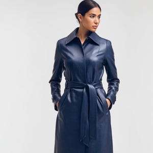 May include: A long, navy blue leather trench coat with a belted waist and a classic collar. The coat features long sleeves and pockets. The model is wearing the coat, with hands in the pockets, against a white background.