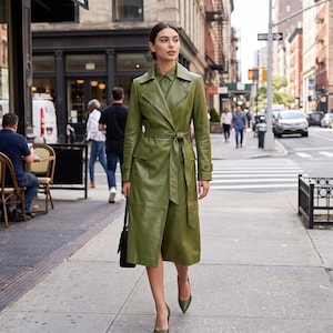 May include: A long, olive green leather trench coat featuring a wide collar, long sleeves with buckled cuffs, and a matching tie belt at the waist. The coat has two front flap pockets and is worn over a button-down shirt, paired with pointed-toe heels.