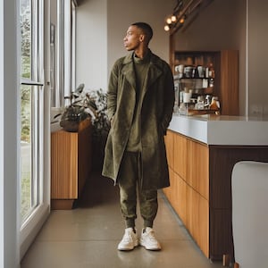 Pode incluir: Um trench coat de camurça verde oliva, gola alta e calças de jogging a condizer. O traje é combinado com tênis brancos. A pessoa está perto de uma janela e de armários de madeira.