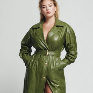 Può includere: Un lungo trench coat verde oliva con cintura e bottoni dorati. Il cappotto ha un classico colletto a revers ed è realizzato in un materiale lucido simile alla pelle. La modella indossa il cappotto con le mani in tasca.