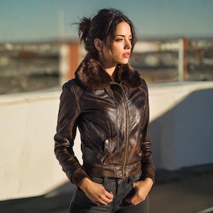 K&ouml;nnte beinhalten: Braune Leder-Bomberjacke mit Kunstfellkragen. Die Jacke hat einen Rei&szlig;verschluss, zwei Vordertaschen und gerippte B&uuml;ndchen. Das Model tr&auml;gt die Jacke mit dunkelgrauen Jeans.
