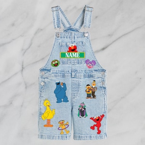 May include: Light blue denim dungarees with Sesame Street character patches. The dungarees have adjustable straps and a front pocket. The name "NAME" is printed on a green label above the chest pocket. Various Sesame Street characters are displayed on the front.