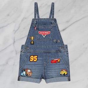 May include: Blue denim overall shorts with various embroidered patches. Patches include a trophy, checkered flags, the number 95, and cartoon car characters. The pocket features the text "NAME".