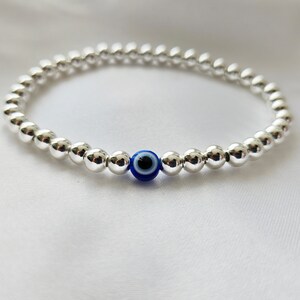 May include: A silver beaded bracelet with a blue and black evil eye charm. The bracelet is made of small, round, shiny silver beads. The evil eye charm is centered on the bracelet and is a deep blue color with a black center.
