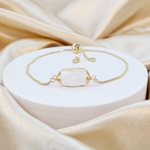May include: A gold-toned bracelet with a rectangular, faceted, light-colored stone at its center. The adjustable chain bracelet is displayed on a white pedestal against a backdrop of draped, shimmering fabric.