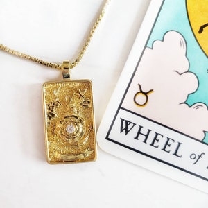 May include: A gold-colored rectangular pendant necklace with a small diamond in the center. The pendant has intricate details and is next to a tarot card that says "WHEEL of ..." and features a Taurus symbol.