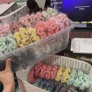 May include: A clear plastic container and a white basket filled with colorful scrunchies in various shades of pink, red, yellow, green, and blue. Each scrunchie is individually packaged. The scrunchies are likely handmade and are a popular hair accessory.