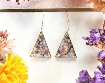 Pode incluir: Brincos triangulares com ganchos dourados. Os brincos apresentam uma resina transparente com uma mistura colorida de flores secas e glitter. Pequenas contas brancas são colocadas nos cantos inferiores. Os brincos são exibidos com flores secas em tons de rosa, roxo e amarelo.
