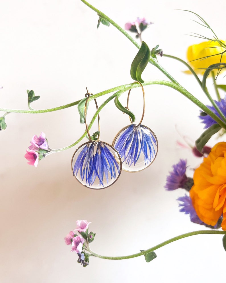 Pode incluir: Um par de brincos de argola dourados com detalhes de flores azuis prensadas. Os brincos est&atilde;o pendurados em um caule verde com flores.