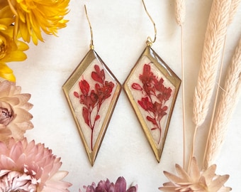 Pode incluir: Um par de brincos em forma de diamante com molduras douradas. Cada brinco apresenta um arranjo floral vermelho encapsulado em resina transparente. Os brincos são exibidos com flores secas em tons de amarelo, rosa e bege.